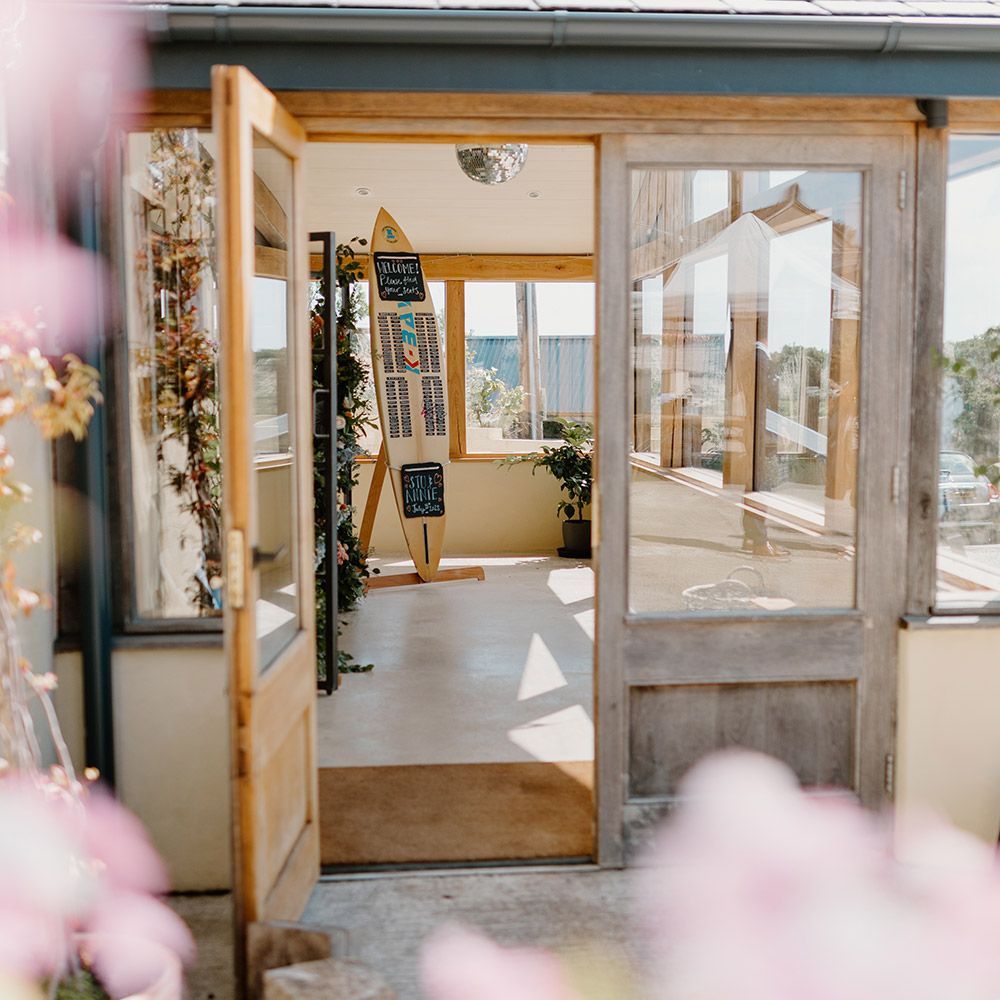 Open doors to a bright room with a surfboard display and foliage.