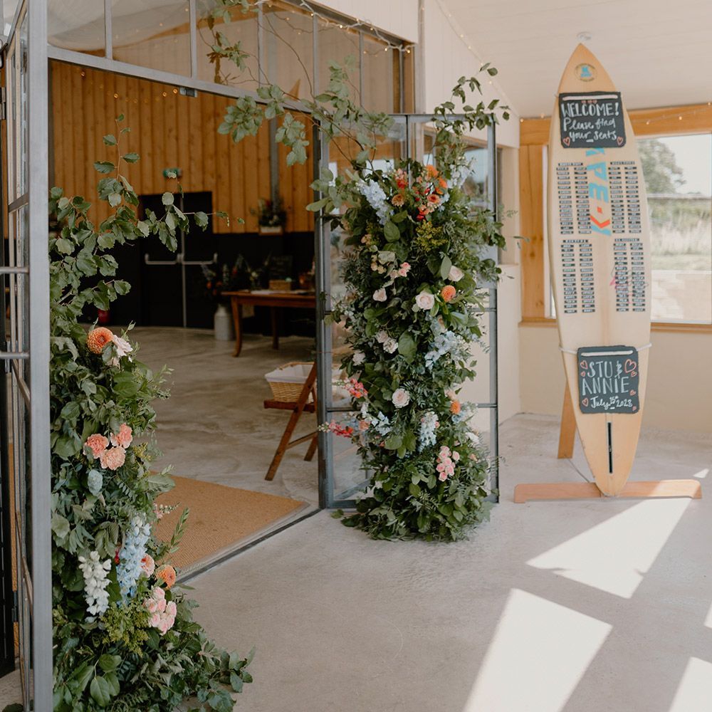 Wedding reception entrance with floral arch, surfboard seating chart, and open doors.