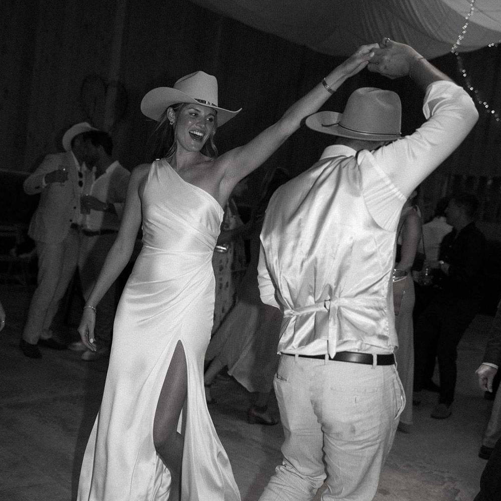 Woman in white gown and cowboy hat dances with a man in a vest and hat at a party.