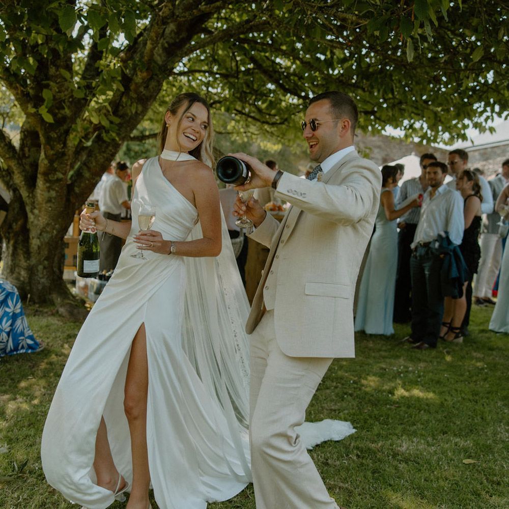 Couple in formal attire celebrating outdoors; man pours champagne, woman smiles, green grass and trees.