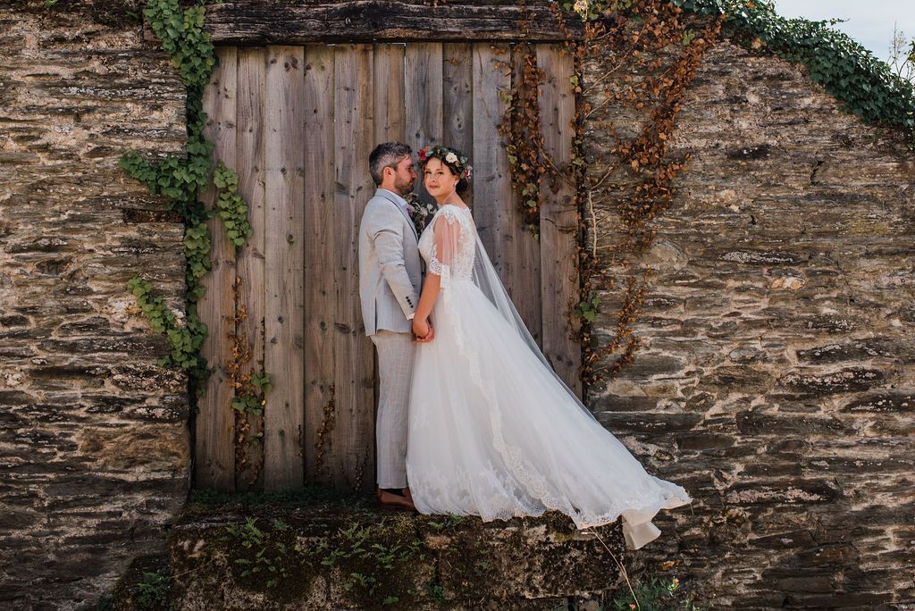 Couple in wedding attire hold hands by a wooden door in a stone building.