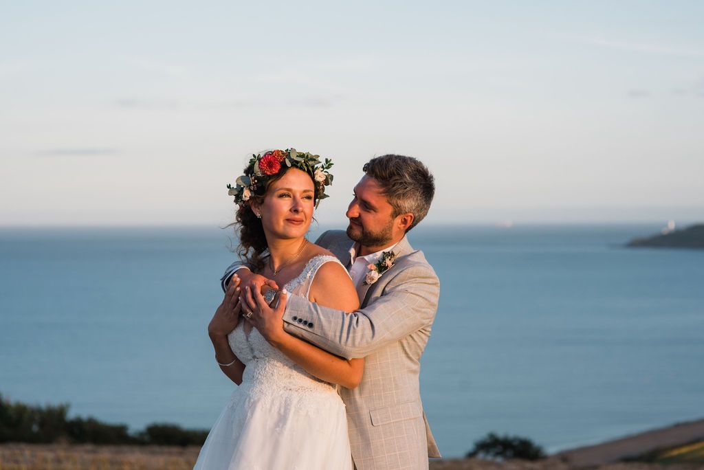 Bride and groom embrace on a hillside overlooking the ocean. The bride wears a floral crown, groom in a suit.