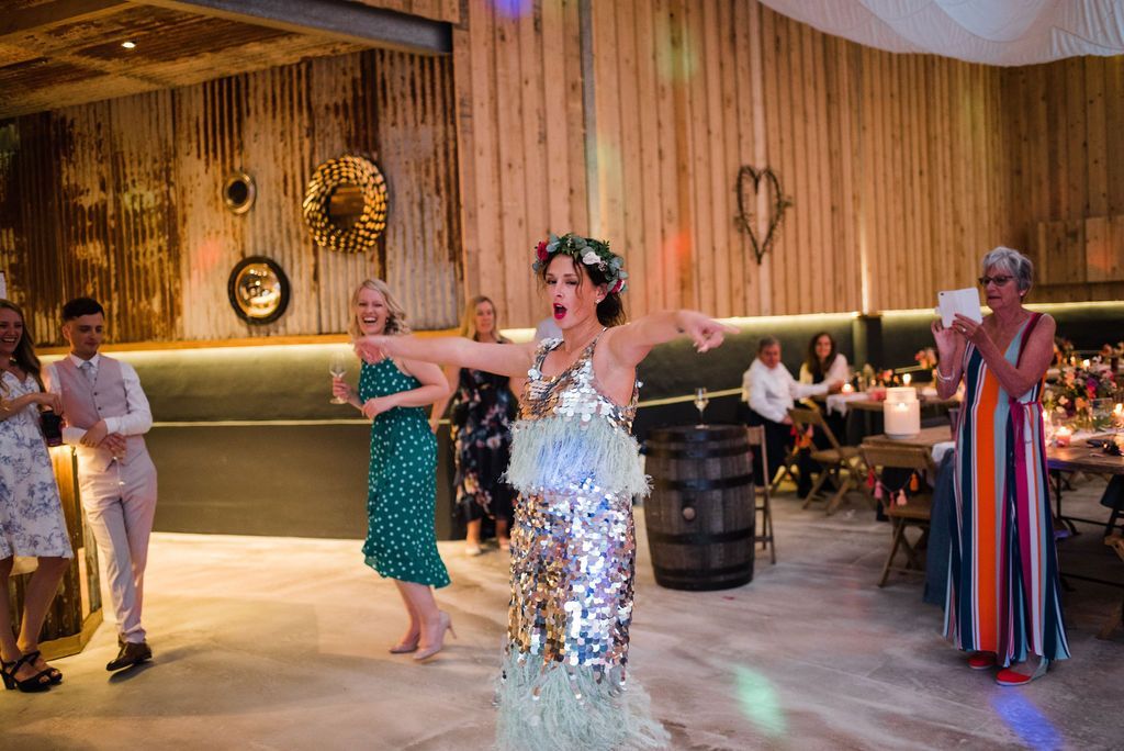 Person in sequined outfit performing at an event, arms outstretched. Guests watch, some with drinks. Wooden interior.