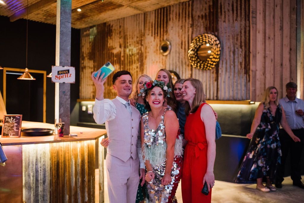 Group of people taking a selfie at a wedding reception; rustic, metal-paneled bar in background.