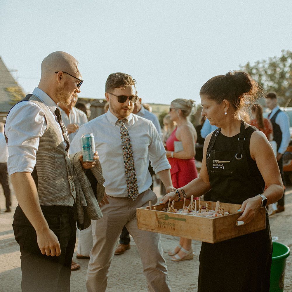 Woman offers appetizers to two men at an outdoor event. Others in background.