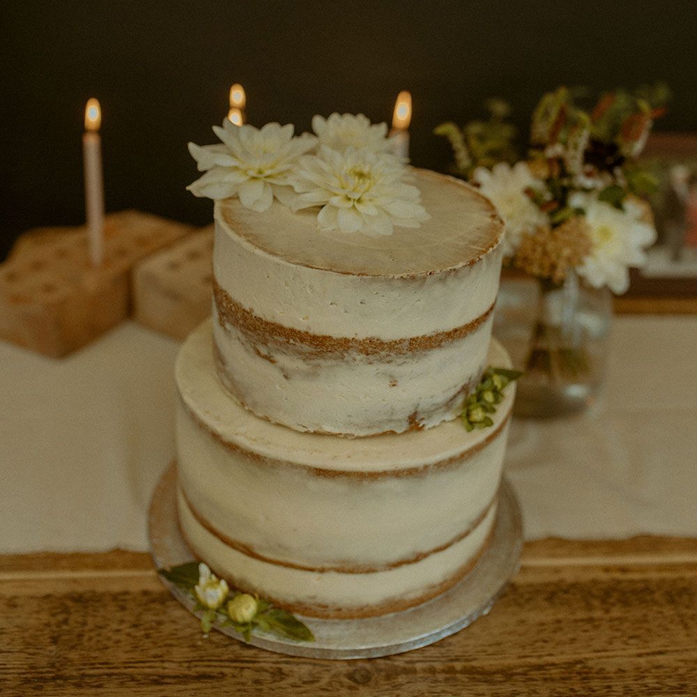 Two-tiered naked wedding cake topped with white flowers, on a wooden table with candles and flowers.