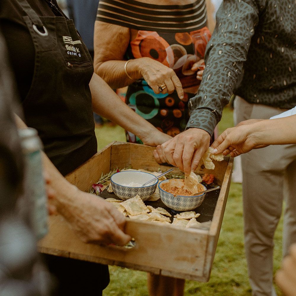 People reaching for snacks from a wooden tray held by a server outdoors. Crackers and dips are visible.