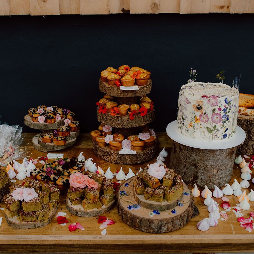 Dessert table with various cakes and pastries on wooden platters, floral decorations, and a rustic setting.