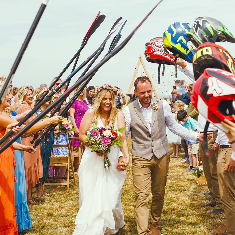 Newlyweds walk between cheering guests holding paddles and helmets outdoors.