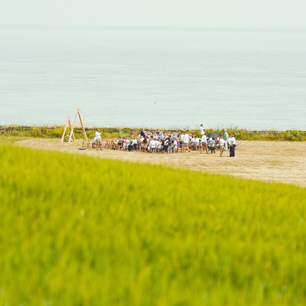 Group of people gathered on a plain near the sea. Tall grass in foreground, blue water and sky in background.