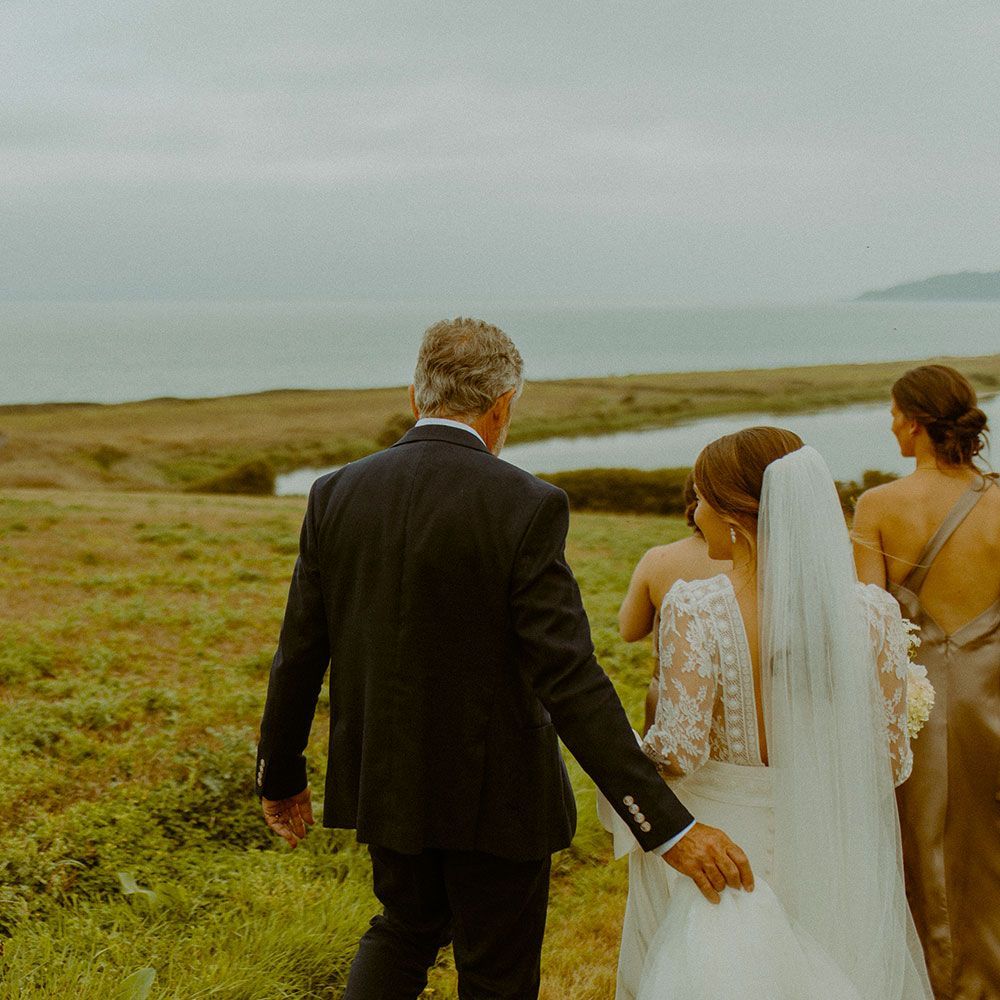 Father walking bride down a grassy hill toward water, bridesmaid follows. Cloudy day.