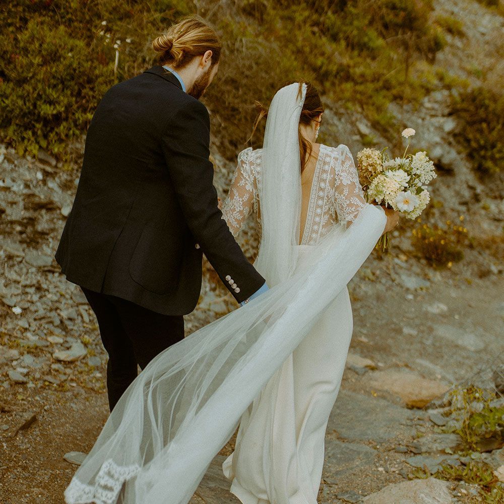 A newlywed couple walks on a rocky path. The bride wears a white dress with a veil, and the groom a black suit.