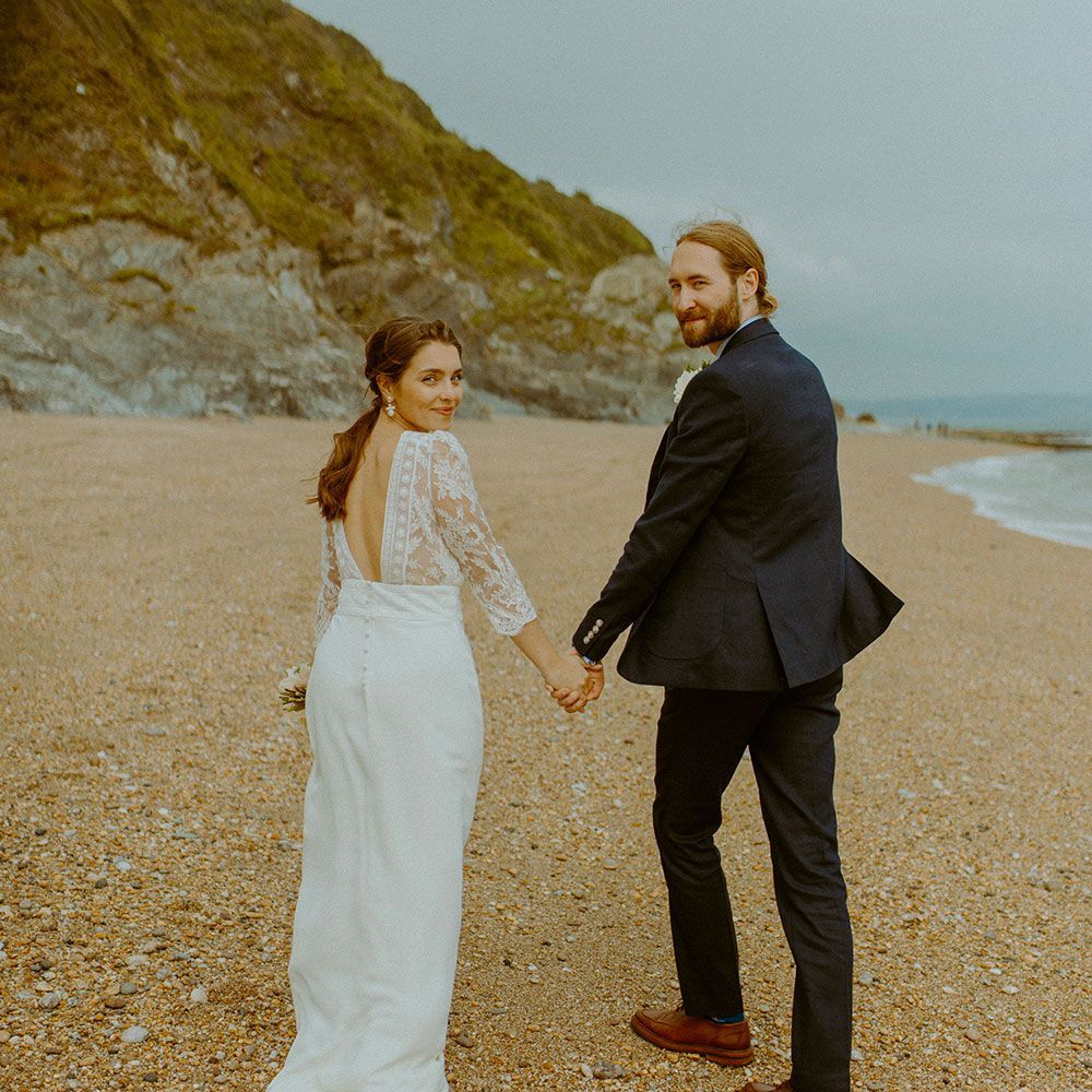Bride and groom holding hands, walking on a beach, looking back. Cliffs and ocean in background.