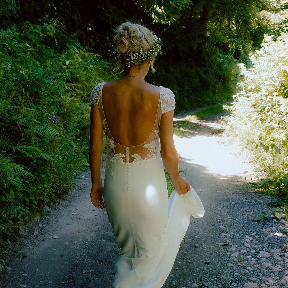 Woman in wedding dress walks on path in forest; backless gown, floral hairpiece.