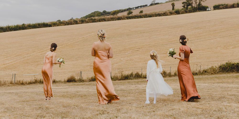 Four women and a young girl walking across a field. The women wear long rust-colored dresses. The girl is in a white dress.