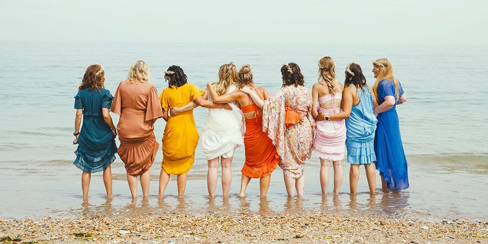 Nine women in colorful dresses stand together in shallow water, arms linked, facing the sea.
