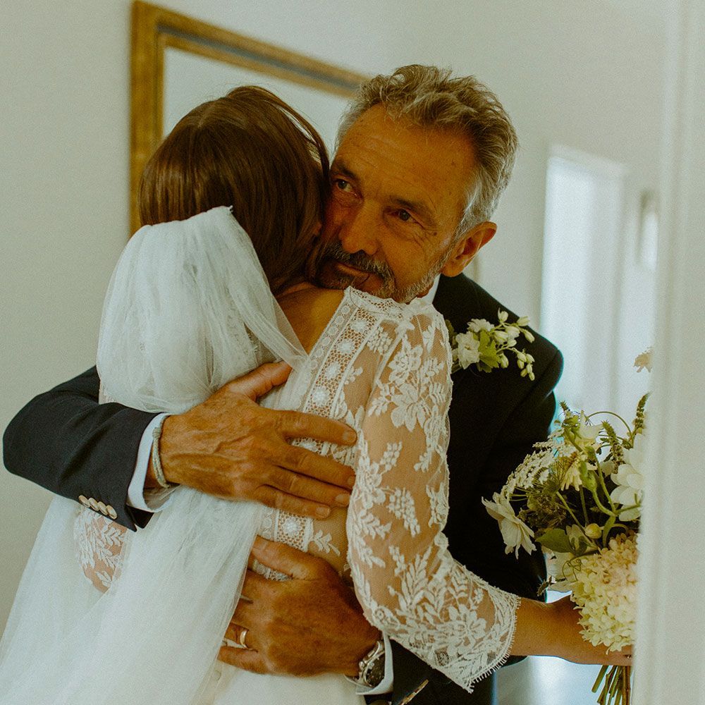 Man hugging woman in a white lace wedding dress, bouquet in hand, near a mirror.