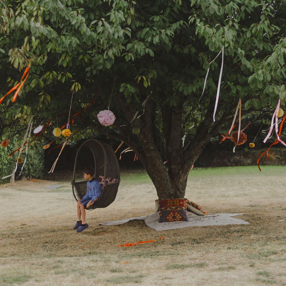 Person sits in a hanging chair under a decorated tree in a yard. Ribbons, decorations.