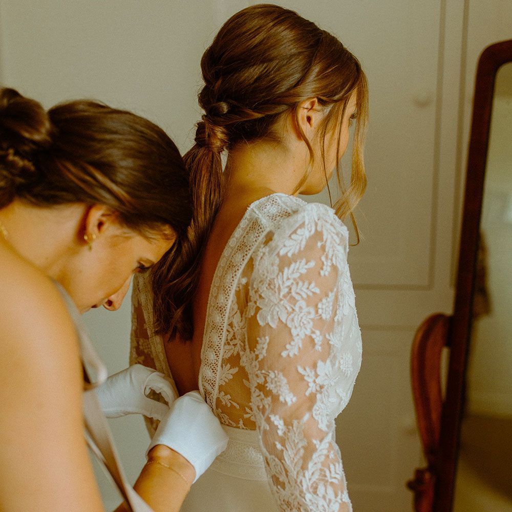 Woman in wedding dress having back of dress adjusted; mirror in background.