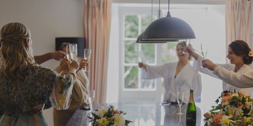 Women in robes toasting champagne, celebrating in a room with a large window and flowers.