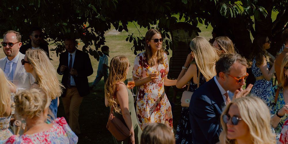 People gathered outdoors, some wearing sunglasses. A woman in a floral dress is talking to others.