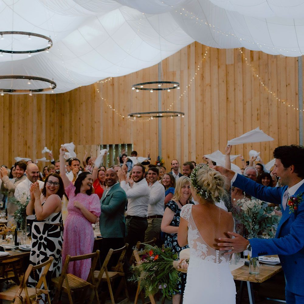 Wedding reception: Bride and groom facing guests, who cheer and wave napkins. Rustic venue with string lights.