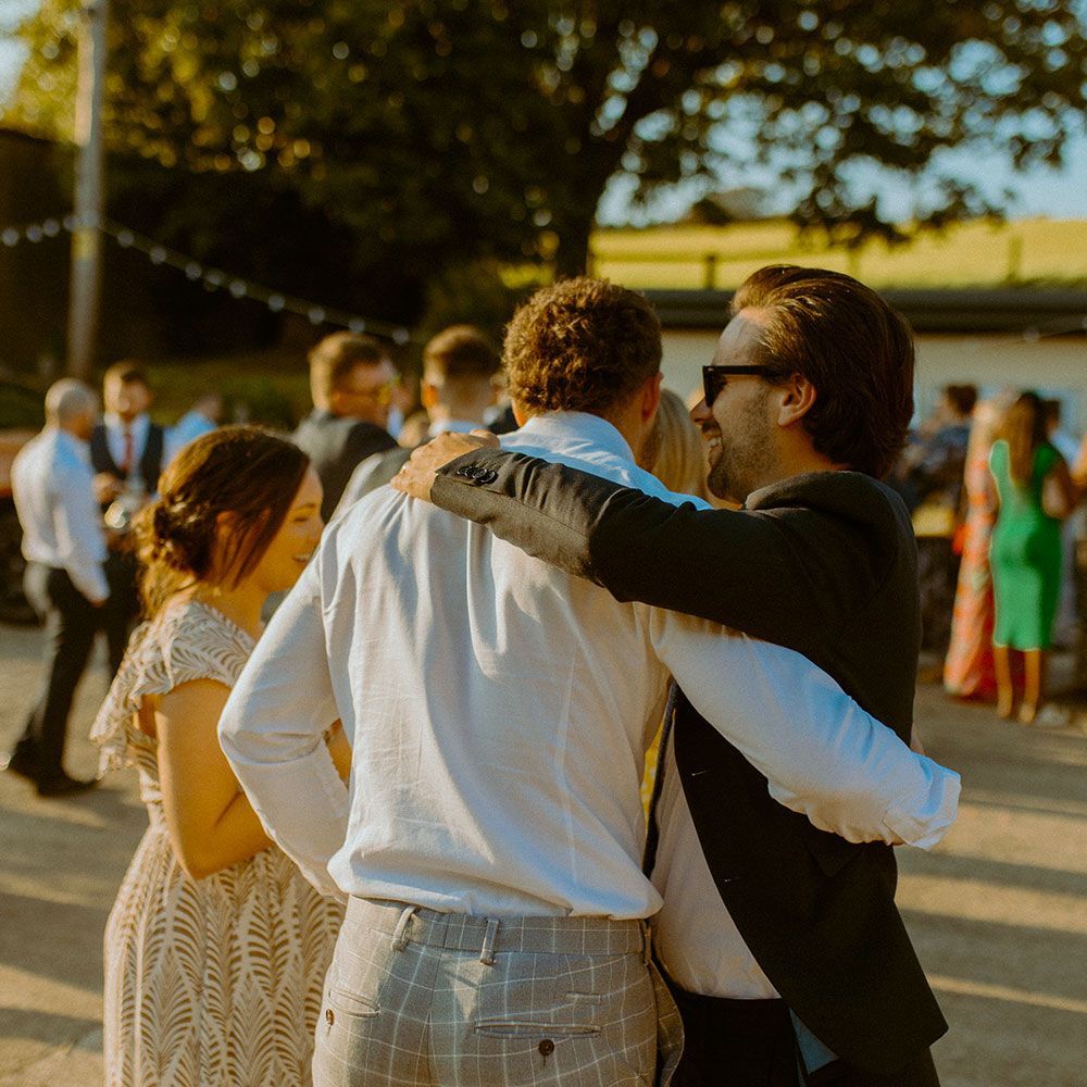 People at an outdoor gathering, two men embracing. Others in formal attire, some with drinks.