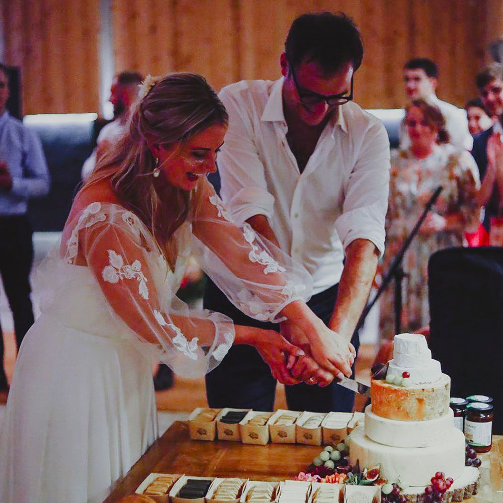Bride and groom cutting cheese wheel cake at wedding reception; indoor setting with guests.