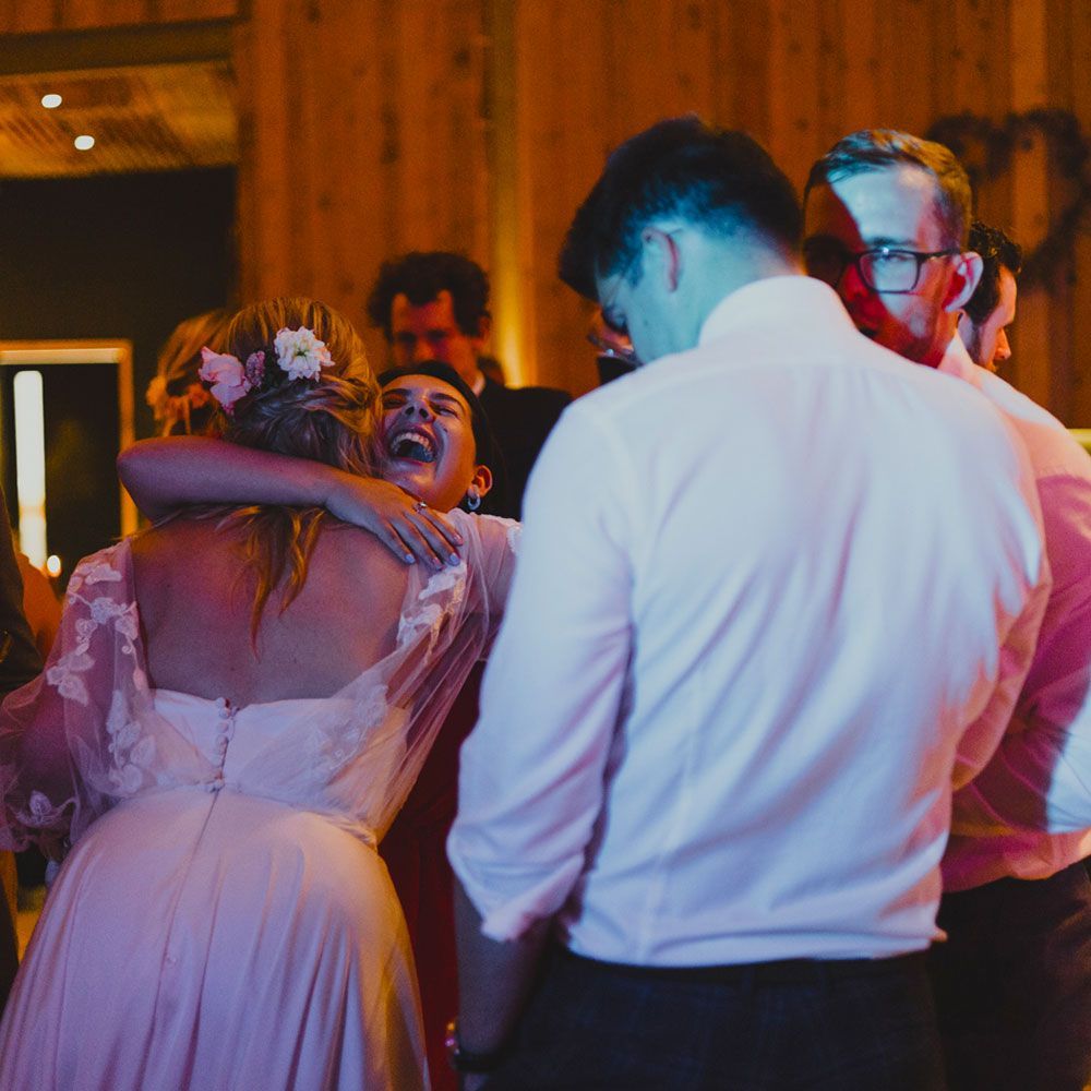 Bride embracing a guest, both smiling; other guests and wood-paneled room in background.