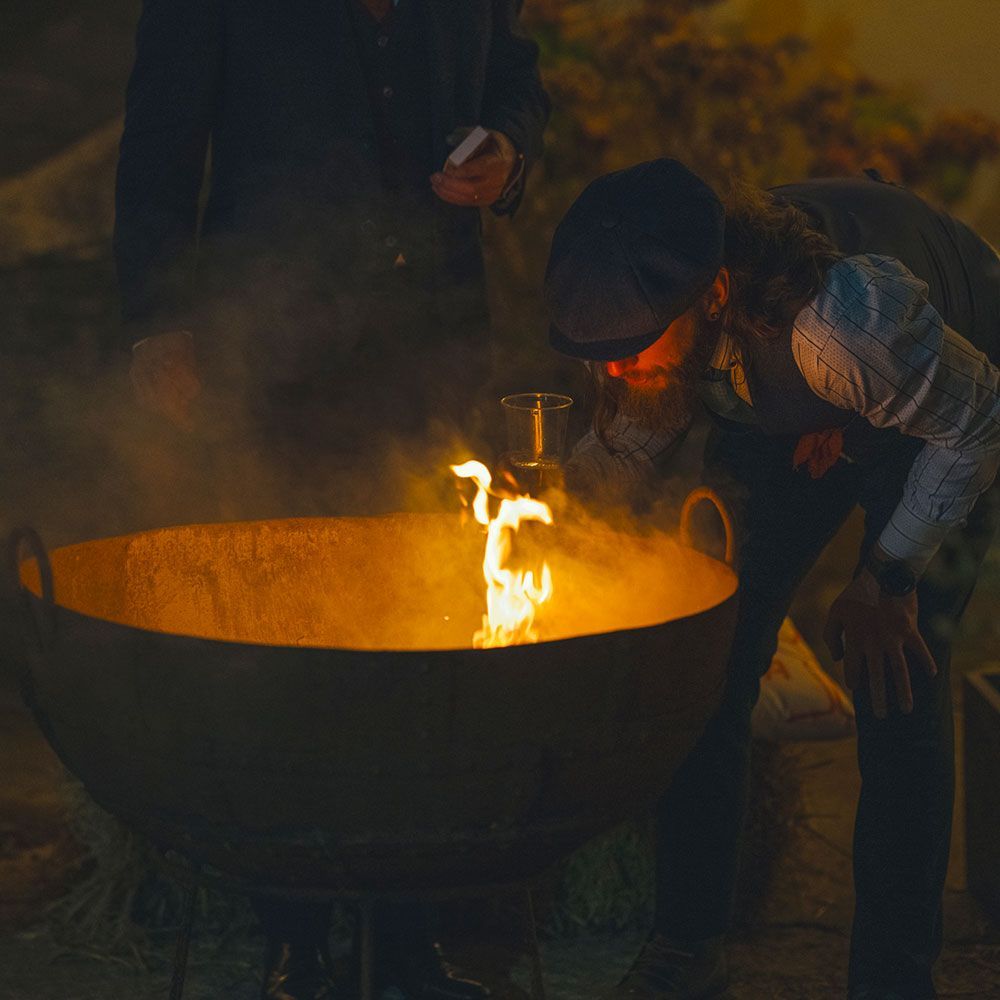 Person ignites something in a large pot with flames, another person nearby. Nighttime.