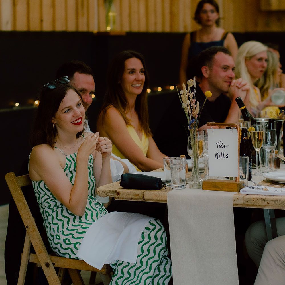 People seated at a table, smiling and clapping. Woman in green and white dress. Table setting with flowers.