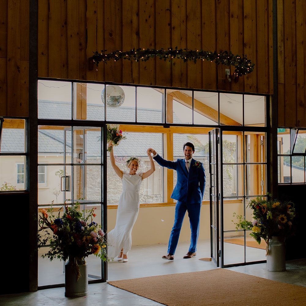 Newlyweds exit a venue, raising arms in celebration. The bride wears a white dress; the groom, a blue suit.