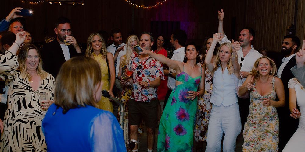 People dancing at an event, some with arms raised and joyful expressions. A person in a blue dress is in the foreground.
