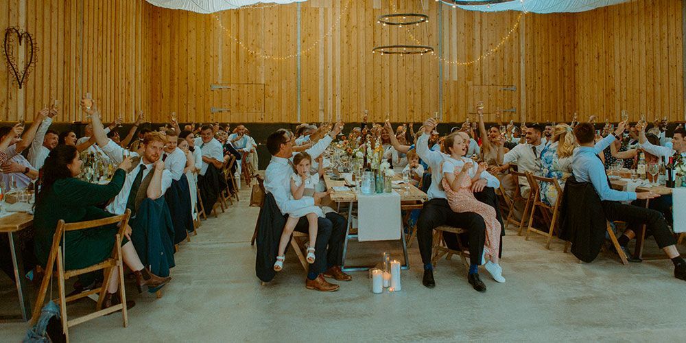 Wedding guests raise their arms in a celebratory toast at a reception, set in a wood-paneled hall.