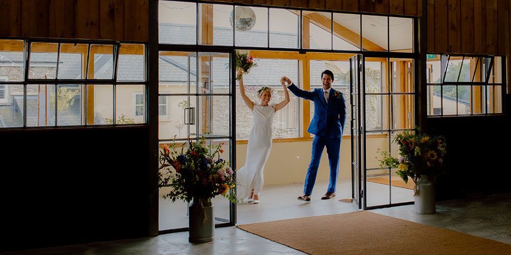 Newlyweds celebrate, exiting a barn door. Bride in white dress, groom in blue suit. Joyful.