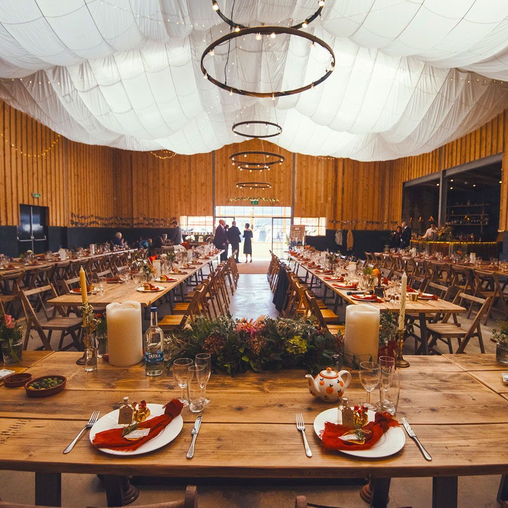 Wedding reception hall with long wooden tables, decorated with flowers and candles, under a draped ceiling.