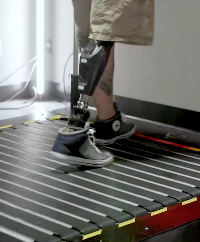 Person walking on a treadmill with a leg brace and support straps attached to the lower leg.