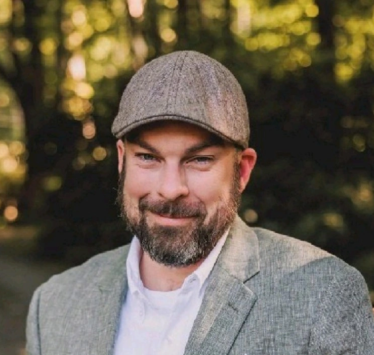 Man in black shirt and flat cap posed against a gray wooden wall