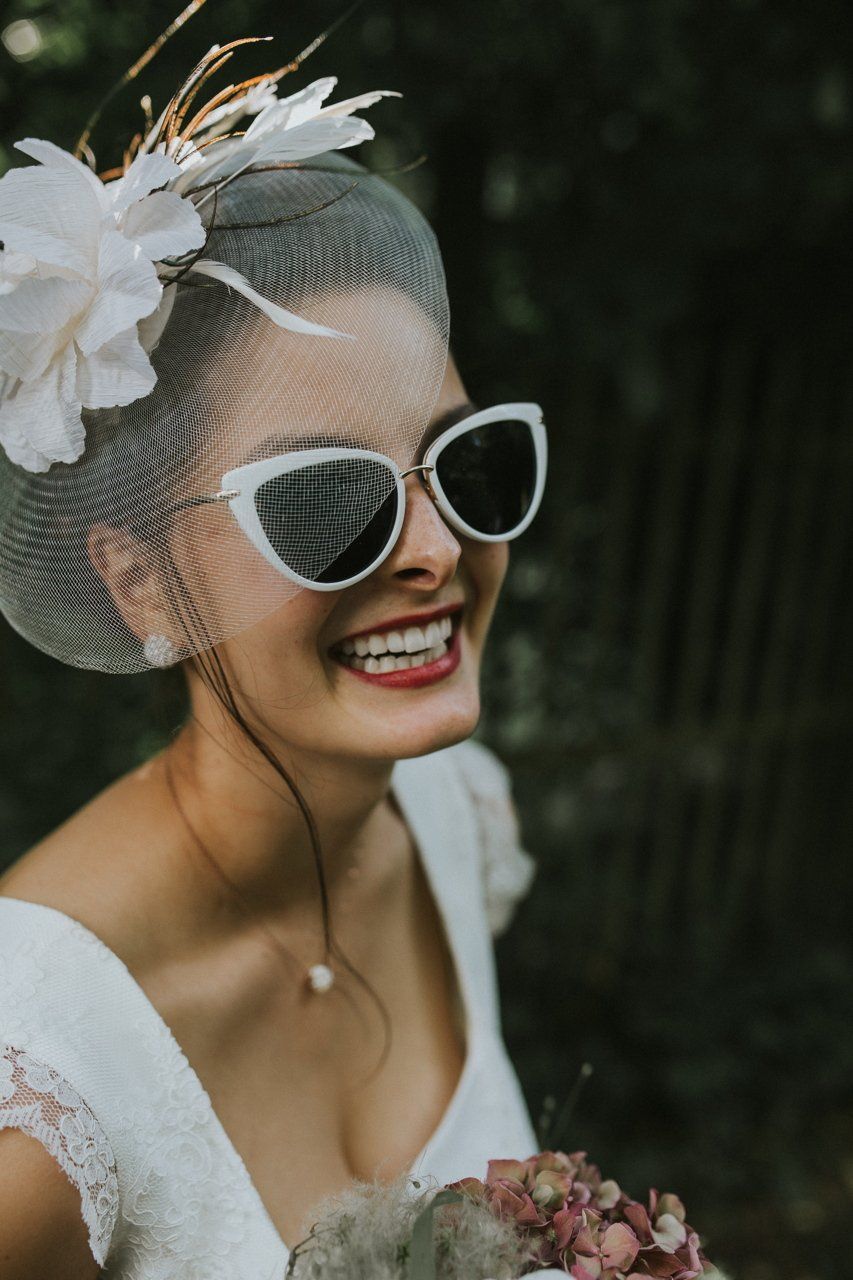 Eine Frau mit Sonnenbrille und Schleier lächelt und hält einen Blumenstrauß in der Hand.