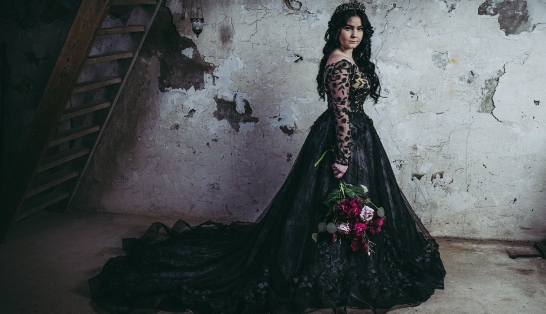 Eine Frau in einem schwarzen Kleid steht mit einem Blumenstrauß in der Hand vor einer Wand.