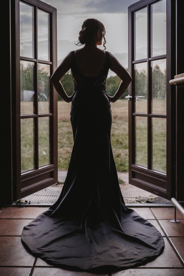 Eine Frau in einem schwarzen Kleid steht vor einem offenen Fenster.