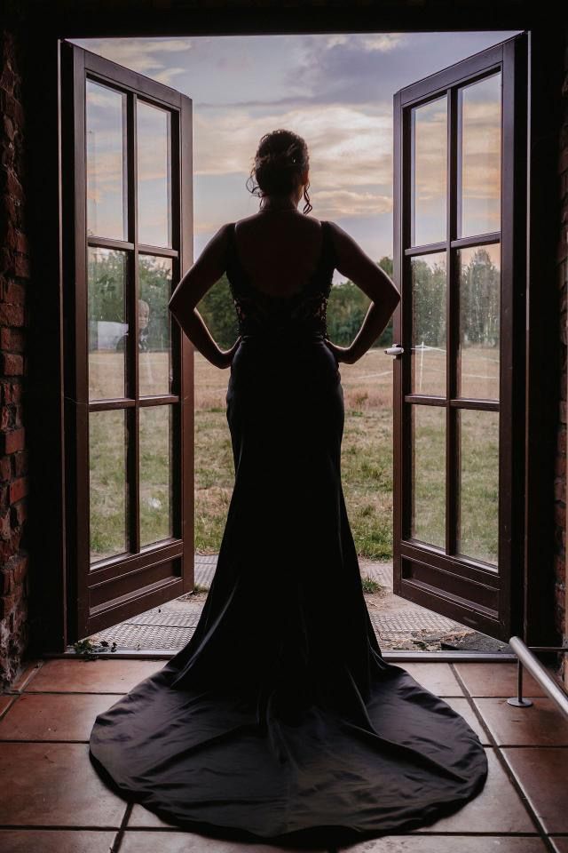 Eine Frau in einem langen schwarzen Kleid steht vor einem offenen Fenster.