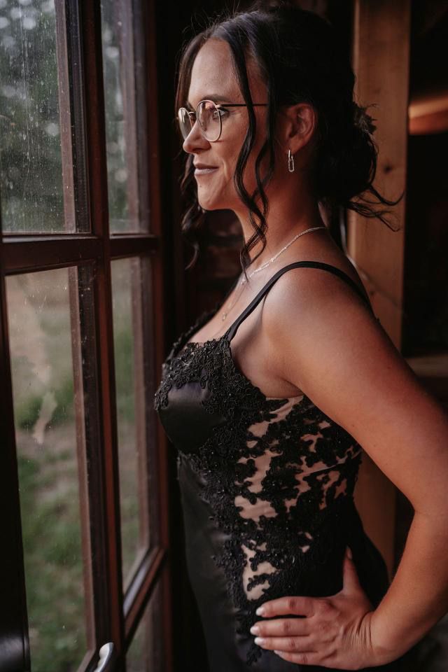 Eine Frau in einem schwarzen Kleid und mit Brille steht vor einem Fenster.