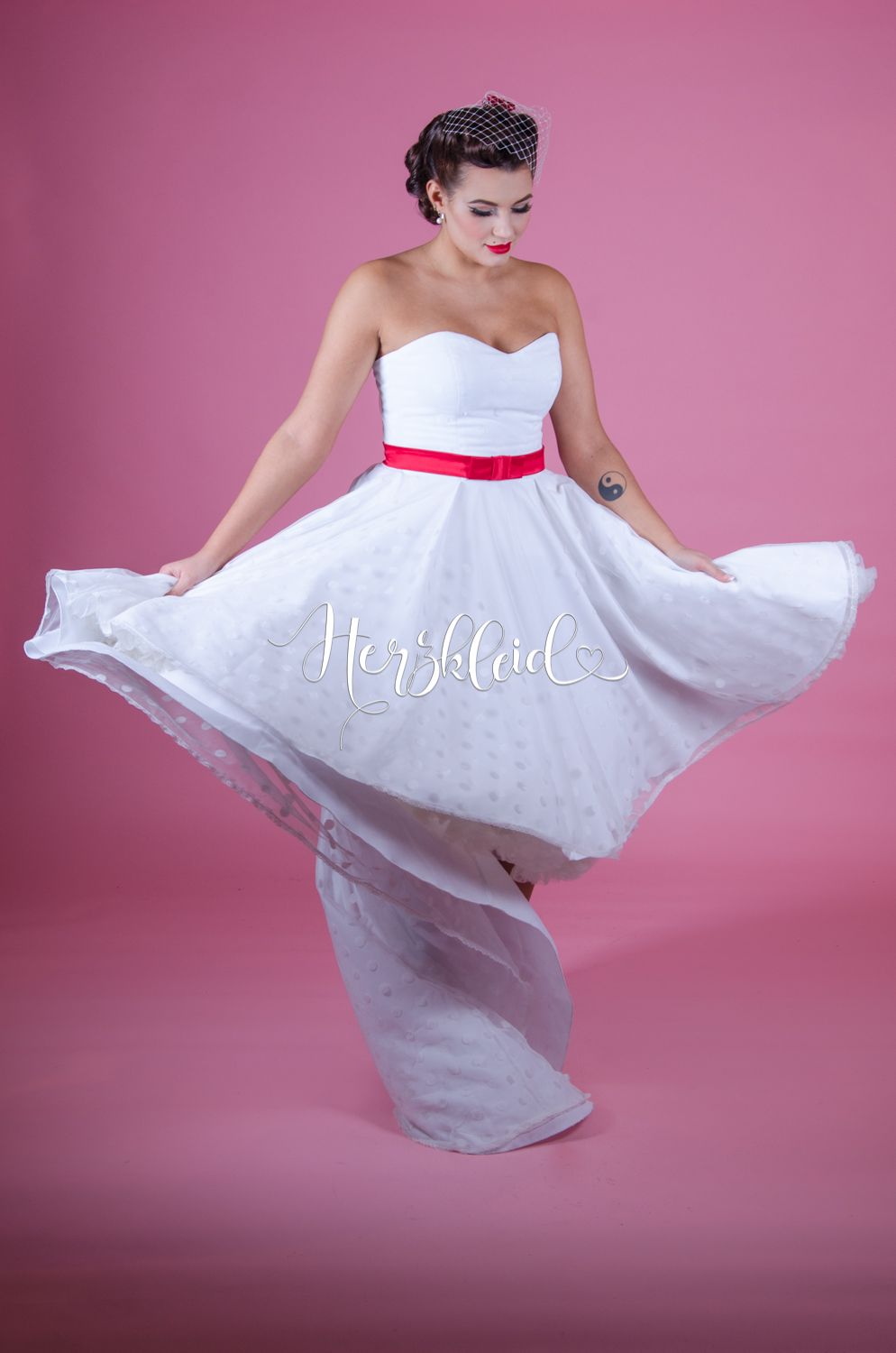 Eine Frau in einem weißen Brautkleid mit roter Schärpe tanzt vor einem rosa Hintergrund.