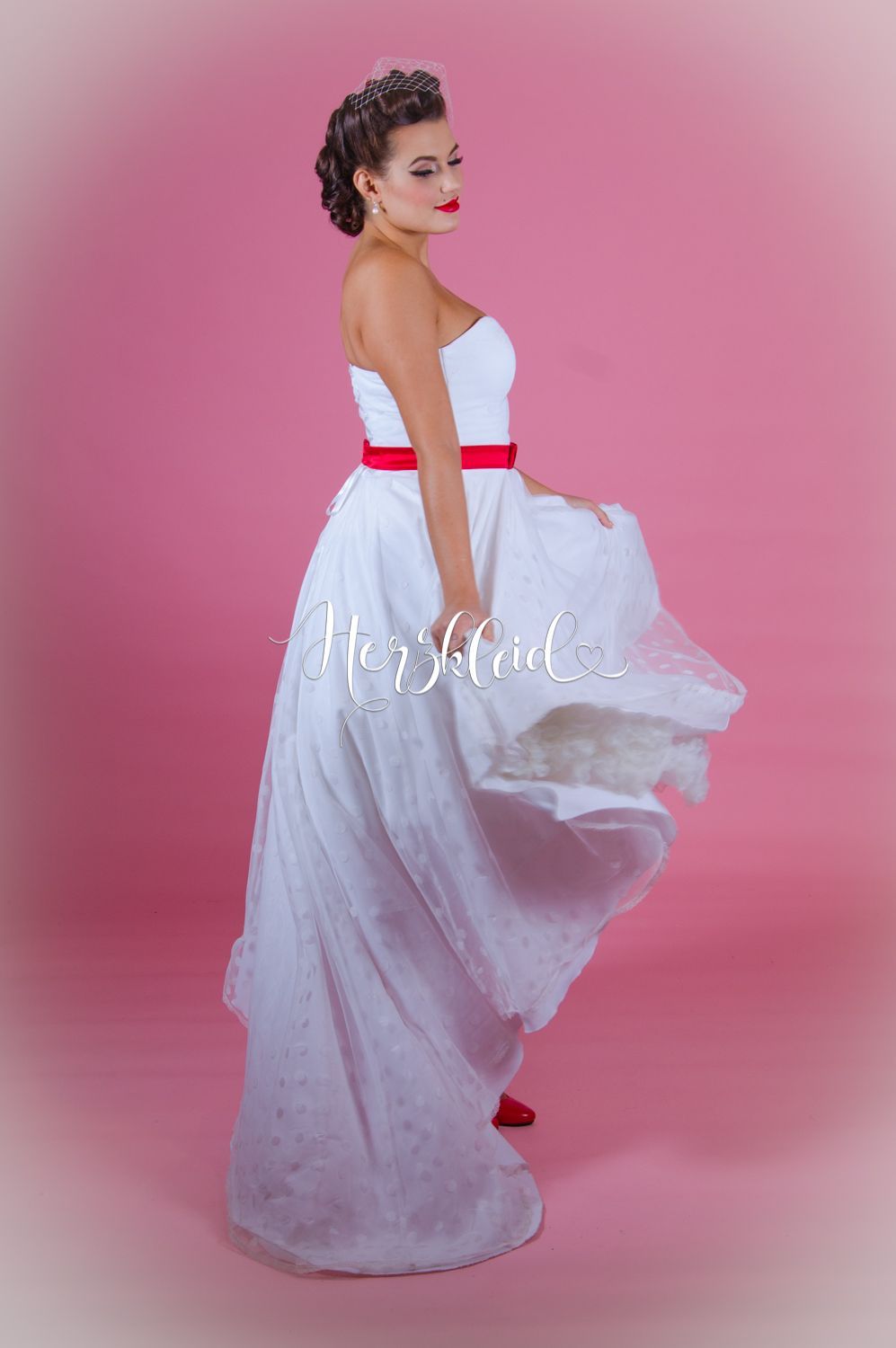 Eine Frau in einem weißen Brautkleid mit rotem Gürtel steht vor einem rosa Hintergrund.