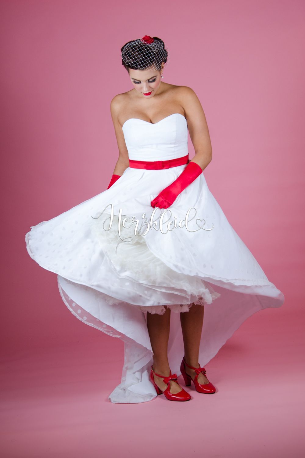 Eine Frau in einem weißen Kleid und roten Handschuhen steht vor einem rosa Hintergrund.