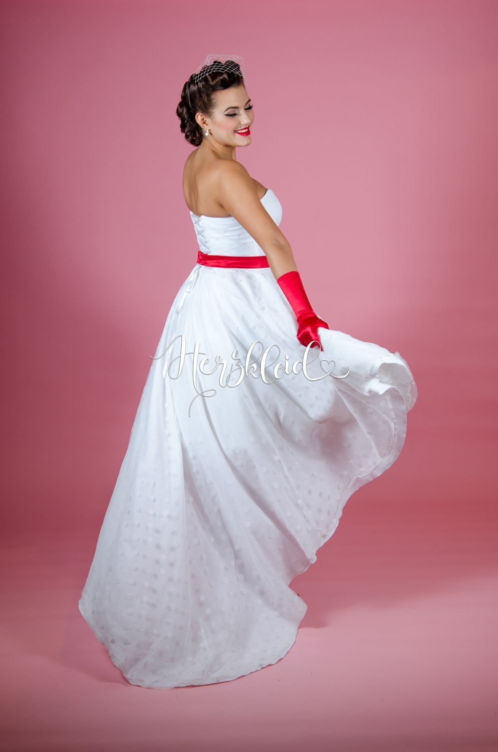 Eine Frau in einem weißen Brautkleid und roten Handschuhen tanzt vor einem rosa Hintergrund.