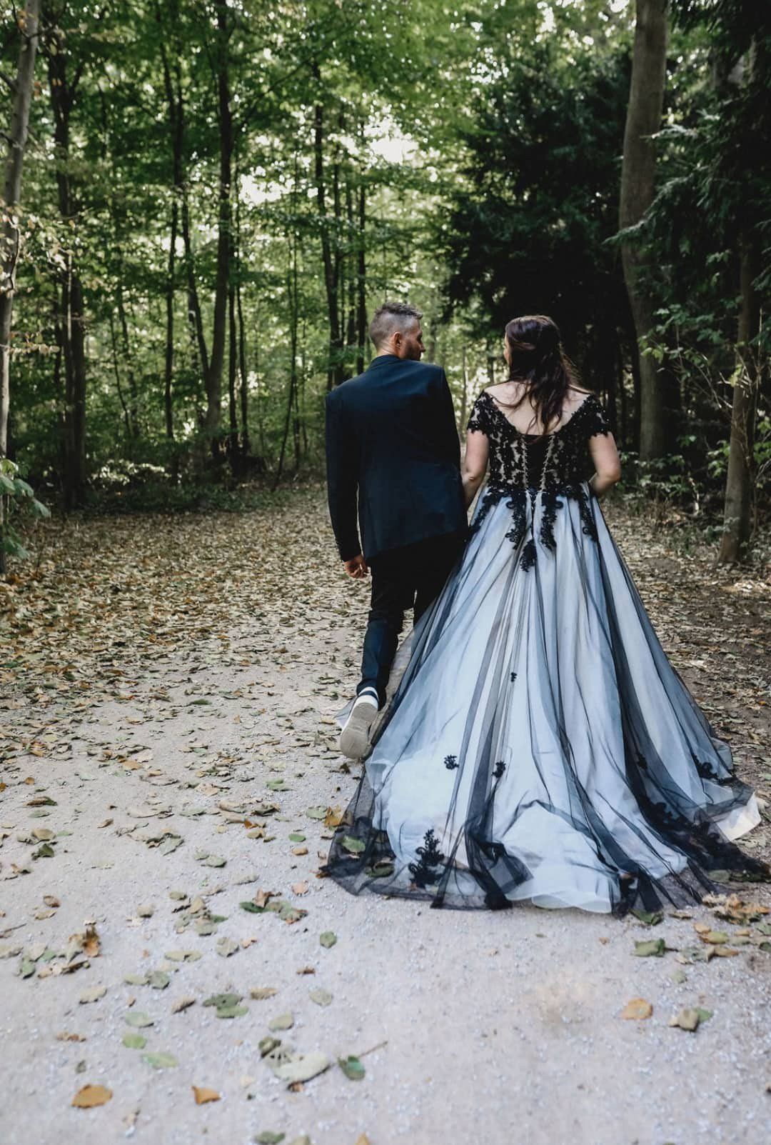 Eine Braut und ein Bräutigam gehen einen Waldweg entlang. Die Braut trägt ein schwarz-weißes Kleid.