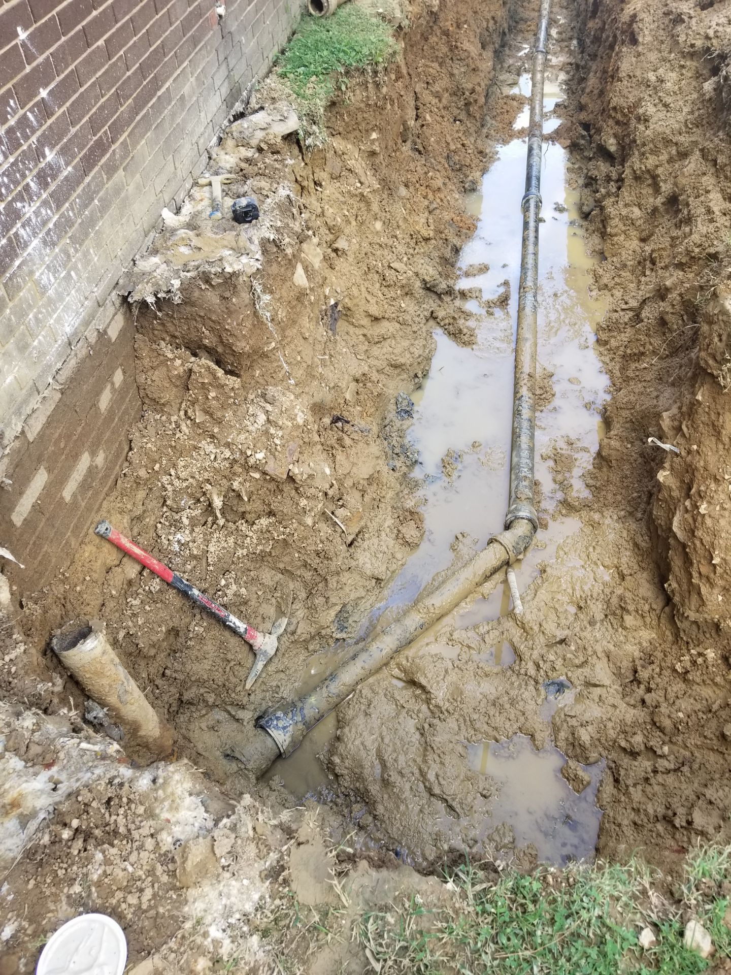Trench with exposed plumbing pipes and tools, next to a brick wall and grass.