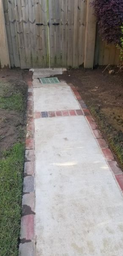 A concrete walkway bordered by red bricks leads to a wooden gate. Lush greenery borders the walkway.